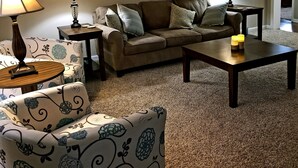 Een woonkamer met een bank, een fauteuil met een patroon, twee bijzettafels met lampen en een salontafel. Boven de bank hangen drie bloemende wanddecoraties.
