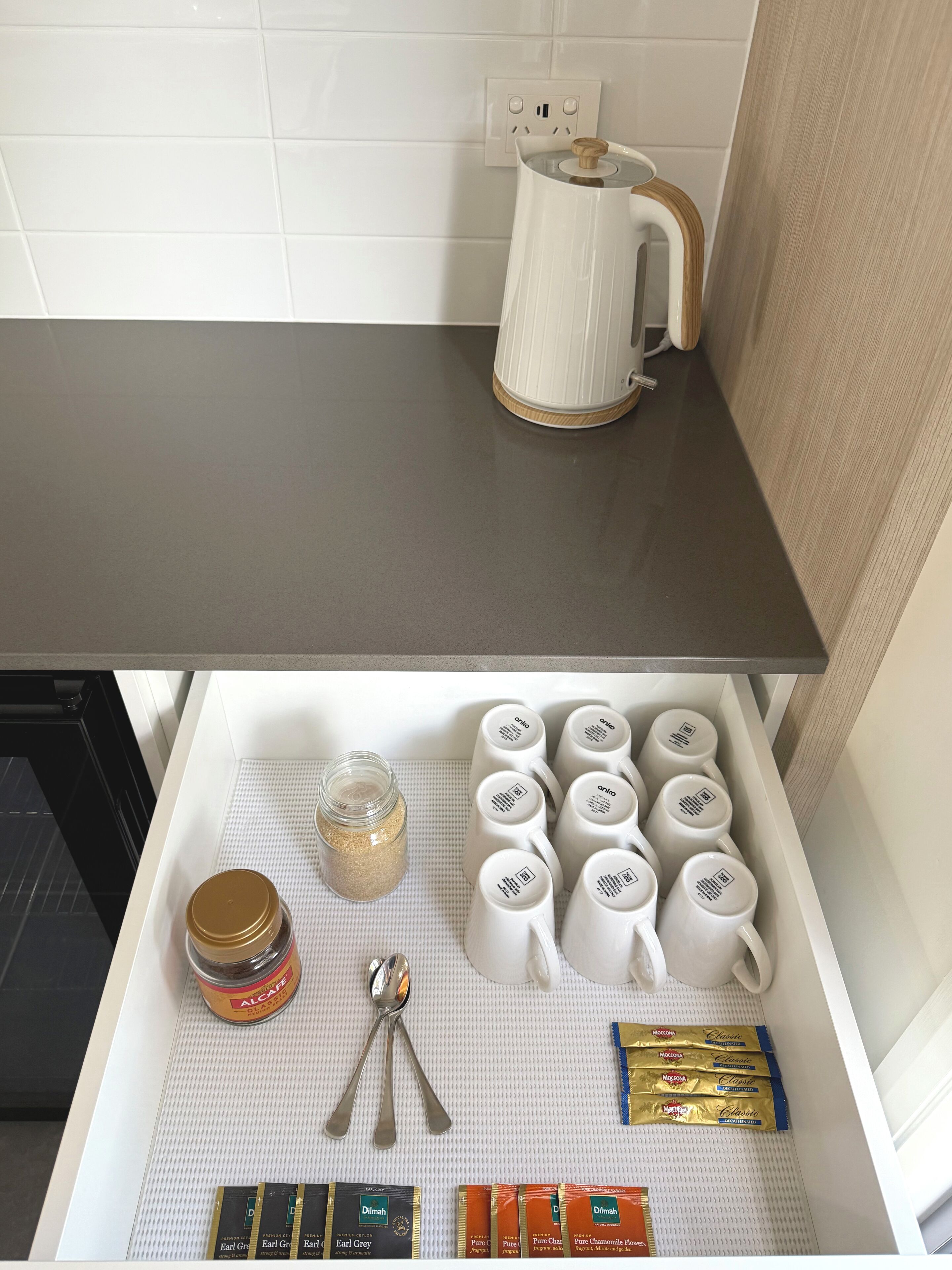 A kitchen counter with a coffee maker and a drawer containing coffee mugs, a jar of coffee, a spoon, and packets of instant coffee.