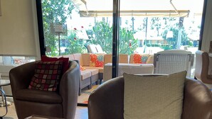 A modern living room with leather sofas and ottomans, a glass door leading outside, and a view of greenery. - Spectacular, South Facing, 4 golf cart, upgraded 2 kings+den with queen  bed. (palm desert)