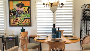 A dining area with a round table set for six, surrounded by wooden chairs with cushioned seats. A chandelier hangs above the table, and a painting is visible on the wall to the left. - Spectacular, South Facing, 4 golf cart, upgraded 2 kings+den with queen  bed. (palm desert)