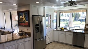 A modern kitchen with white cabinetry, a stainless steel refrigerator, and a large window offering a view of a backyard. - Spectacular, South Facing, 4 golf cart, upgraded 2 kings+den with queen  bed. (palm desert)