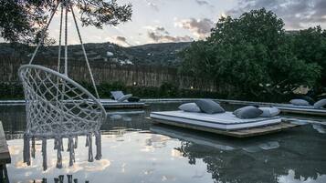 Un área de piscina al aire libre tranquila, con camas flotantes y una silla colgante, rodeada de exuberante vegetación y colinas al fondo.