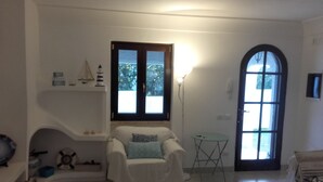Een woonkamer met een witte fauteuil, een bijzettafeltje, een lamp en een raam met uitzicht op bomen.