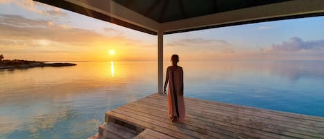 A person standing on a wooden deck overlooking a calm sea at sunset.