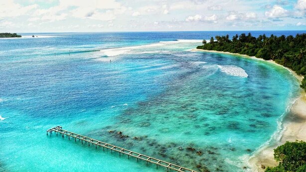 Vista costera con aguas turquesas cristalinas, una playa arenosa y un muelle de madera que se adentra en el mar.