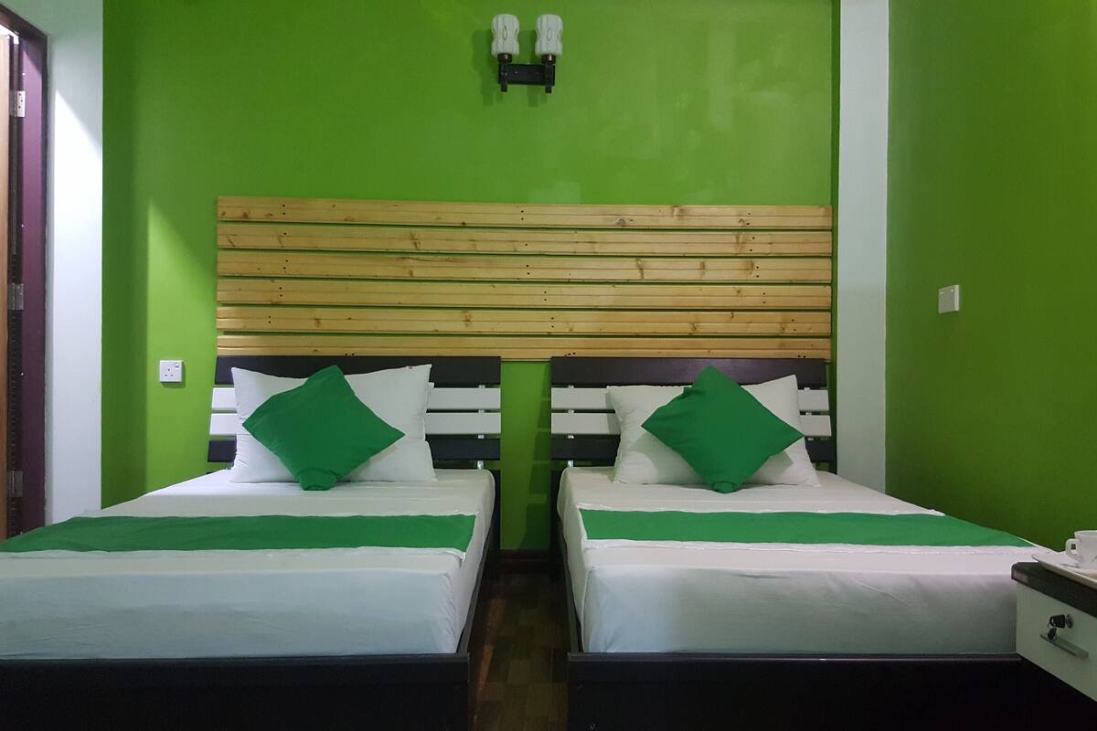 Una habitación con dos camas, cada una con cabecero de madera, ropa blanca de cama y almohadas verdes. A la derecha, hay una mesita con una taza y un plato.