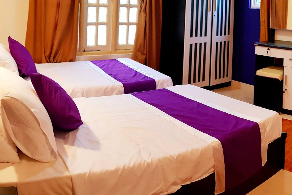 Habitación de hotel con dos camas, cada una con ropa de cama blanca y morada, cabeceros de madera, una ventana con cortinas y un escritorio pequeño con una silla.
