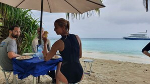 Un montaje para comer frente al mar, con una mesa cubierta por un paño azul, una persona comiendo un plátano y un barco llamado 'EMPEROR VIRGO' al fondo.