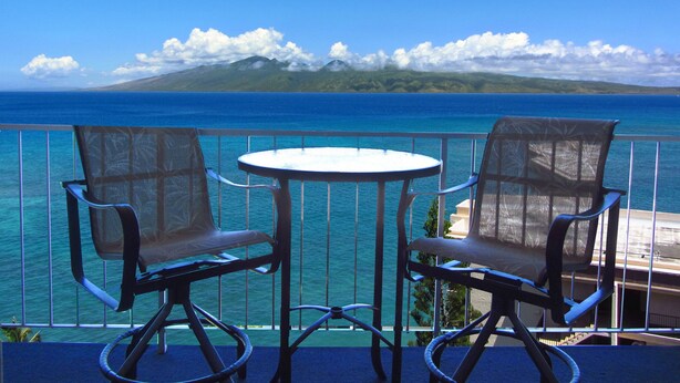 Een balkon met twee stoelen en een tafel, met uitzicht op een heldere blauwe zee en een verafgelegen bergketen.