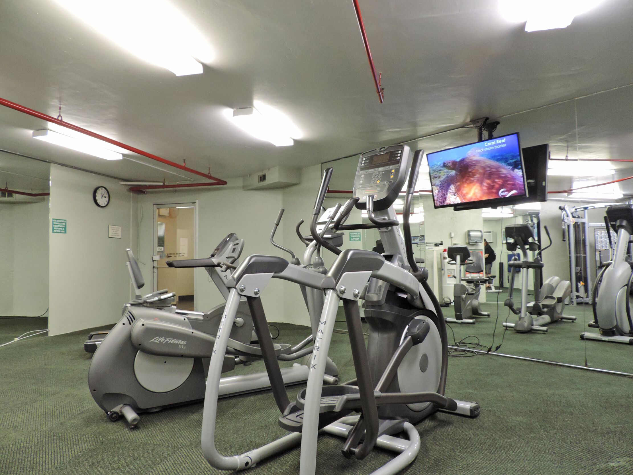 Un gimnasio con equipo de ejercicio, un reloj en la pared y una pantalla de televisión mostrando una imagen de un arrecife de coral.
