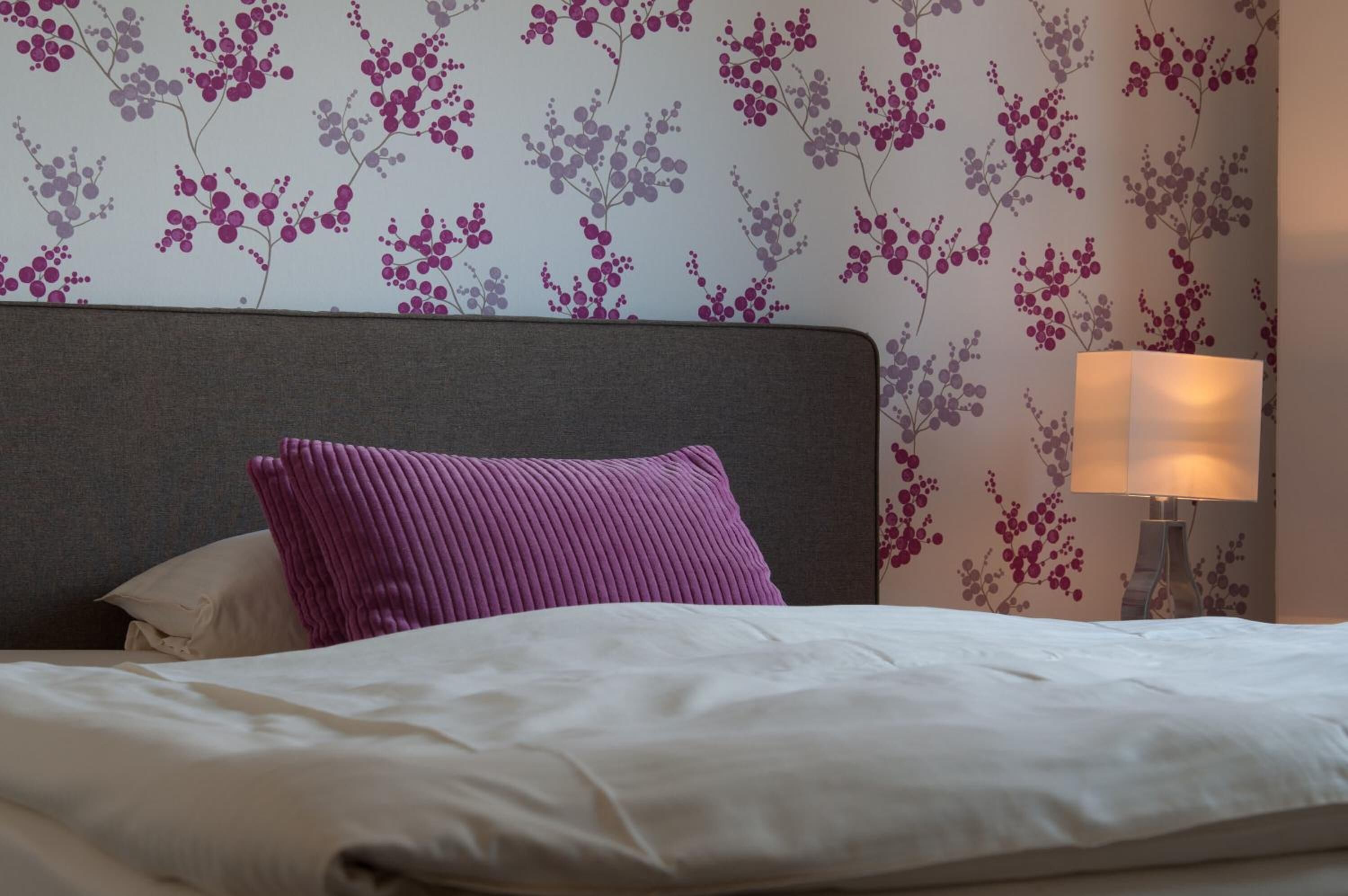 A neatly made bed with white linens and a purple pillow, a grey headboard, and a wall with floral wallpaper.
