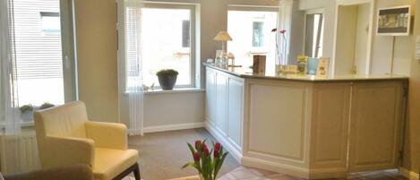 A spacious living room with a sofa, a coffee table with a vase of flowers, a kitchen area with a counter and cabinets, and a large window providing natural light.