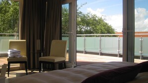Quarto de hotel com cama, poltrona, mesa e varanda com guarda-corpo de vidro.