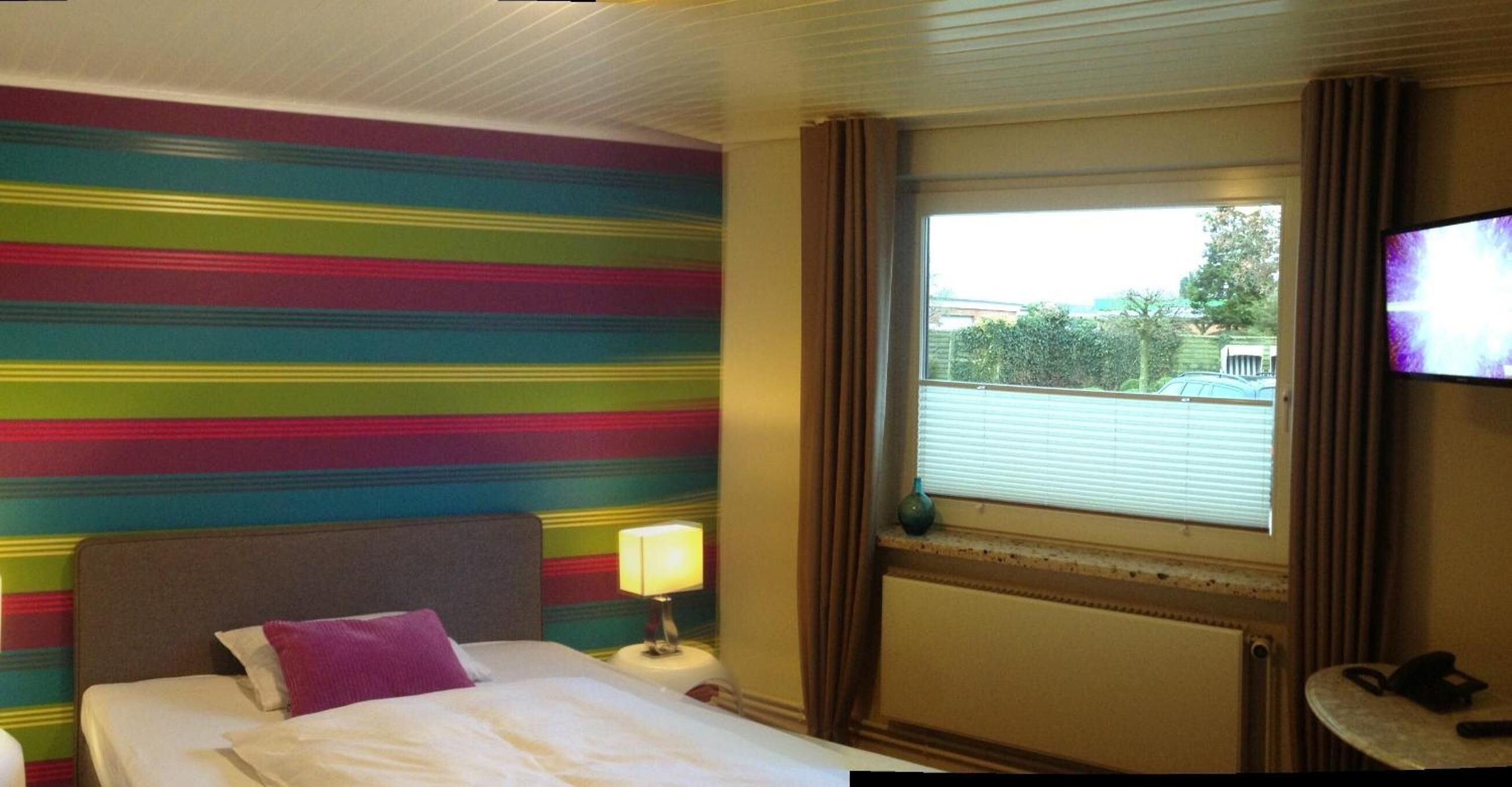 Quarto de hotel com cama, mesa de cabeceira com luminária, janela com cortinas e televisão na parede.