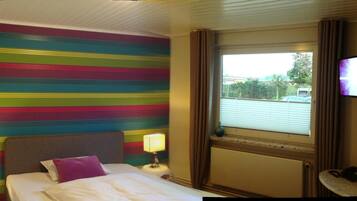 Quarto de hotel com cama, mesa de cabeceira com luminária, janela com cortinas e televisão na parede.