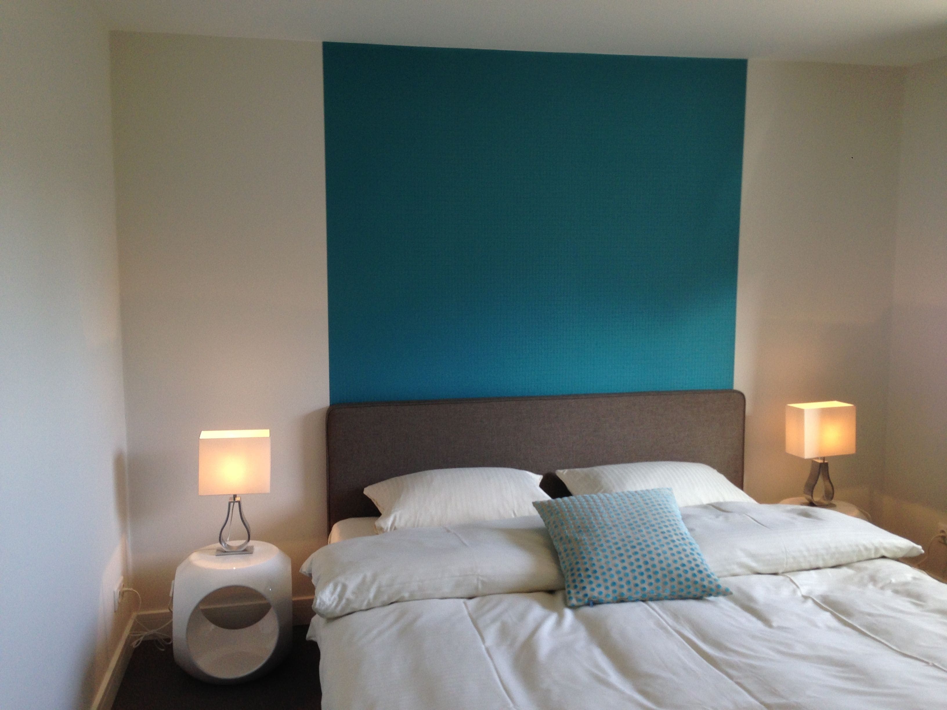 A neatly made bed with white linens and a teal accent pillow, flanked by two bedside lamps on white nightstands.