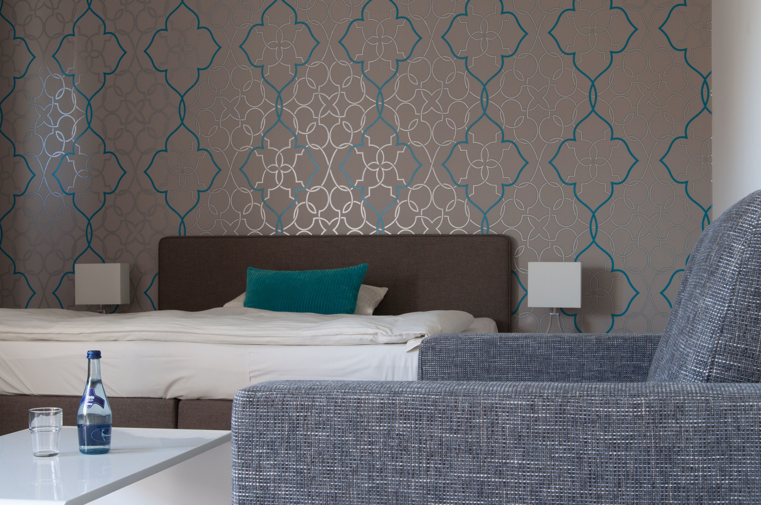 A hotel room with a patterned wall, a bed with white bedding and a teal pillow, two bedside lamps, a grey upholstered armchair, and a small table with a bottle and glass.