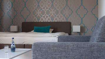 A hotel room with a patterned wall, a bed with white bedding and a teal pillow, two bedside lamps, a grey upholstered armchair, and a small table with a bottle and glass.
