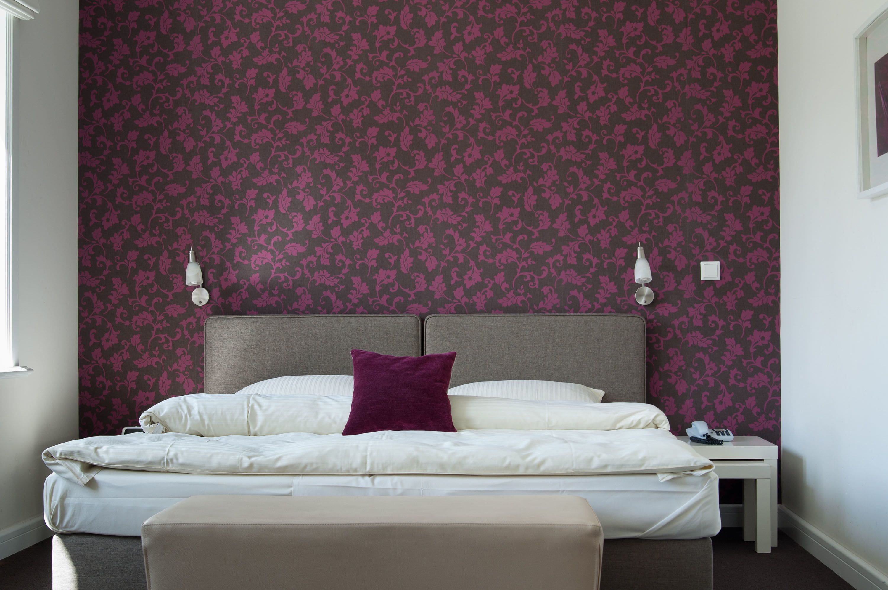 Ein ordentlich bezogenes Bett mit weißen Leinentüchern und einem violetten Kissen, ein Nachttisch mit einer Lampe und eine Wand mit einem Blumenmuster.