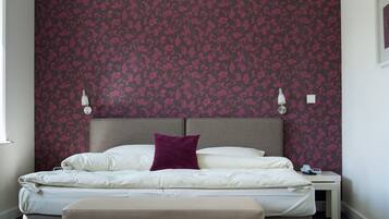Ein ordentlich bezogenes Bett mit weißen Leinentüchern und einem violetten Kissen, ein Nachttisch mit einer Lampe und eine Wand mit einem Blumenmuster.