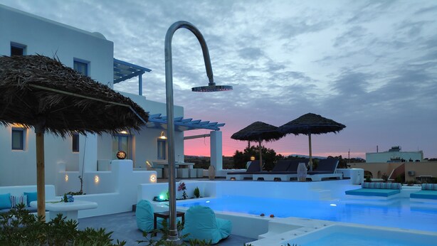 A modern outdoor pool area with lounge chairs and umbrellas, surrounded by white buildings and a clear sky. - Santoxenia Luxury Villa (Santorini)