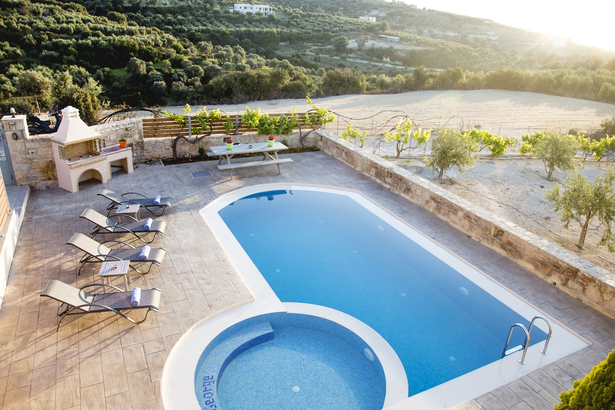 A swimming pool with lounge chairs and a barbecue area, surrounded by a stone wall and a view of the countryside.