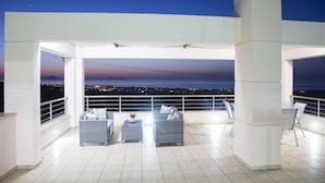 A covered outdoor seating area with wicker furniture and a view of the city at dusk. - Villa Saint George (Rethymno)
