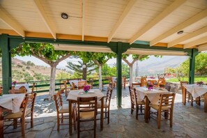 An outdoor patio with stone flooring and wooden tables and chairs, surrounded by greenery and a scenic view.