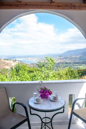A balcony with a table set for two, overlooking a scenic landscape with mountains and a body of water.
