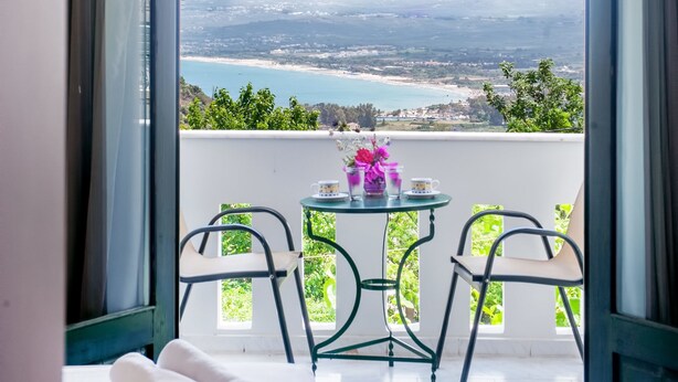 A balcony with a bed, two chairs, a small table with a vase, and a view of a beach and cityscape. - Harma Rooms & Apartments (Apokoronas)