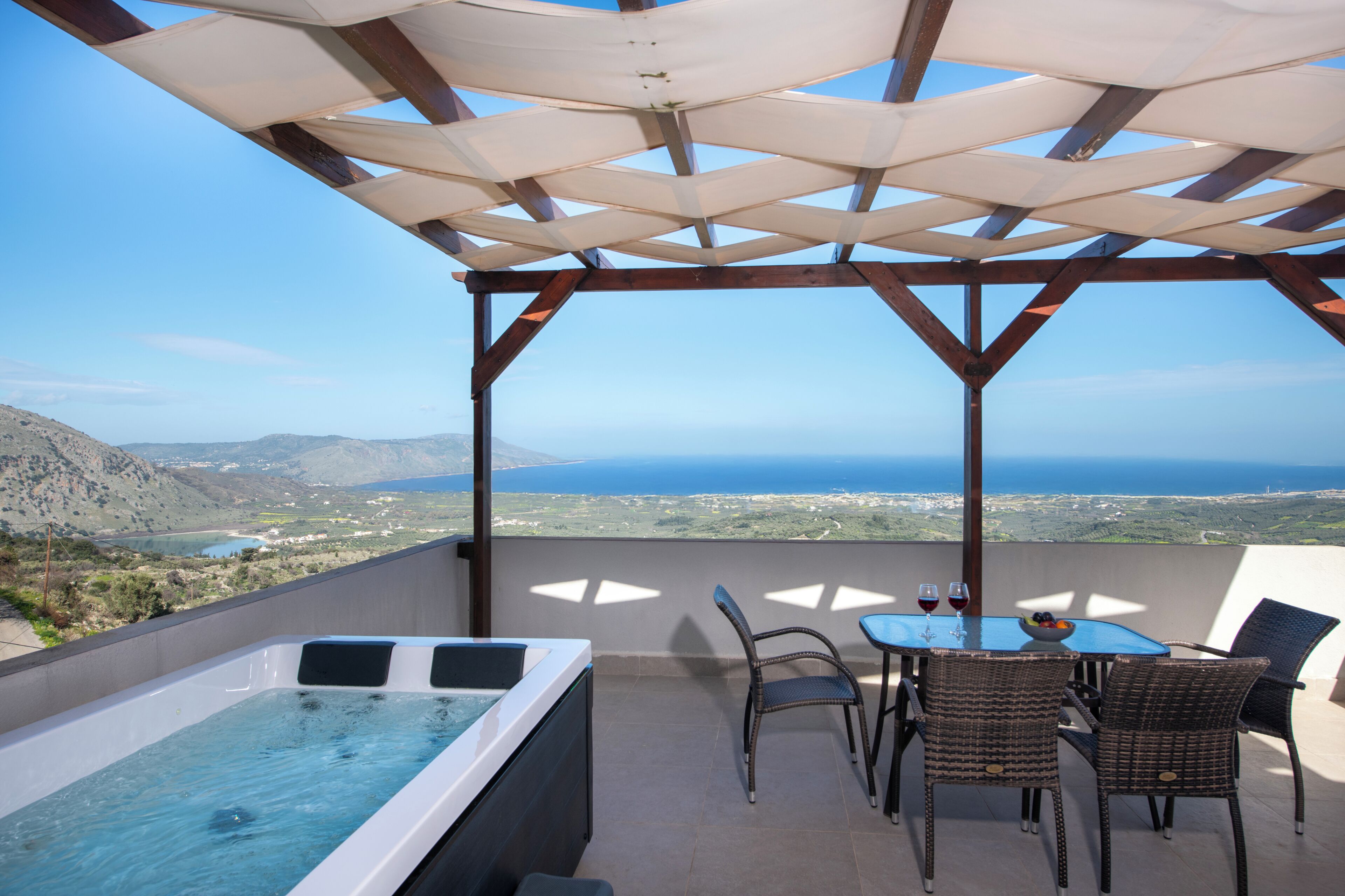 Una terraza en la azotea con un jacuzzi, sillas de mimbre, una mesa con vasos y vistas al océano y las colinas.