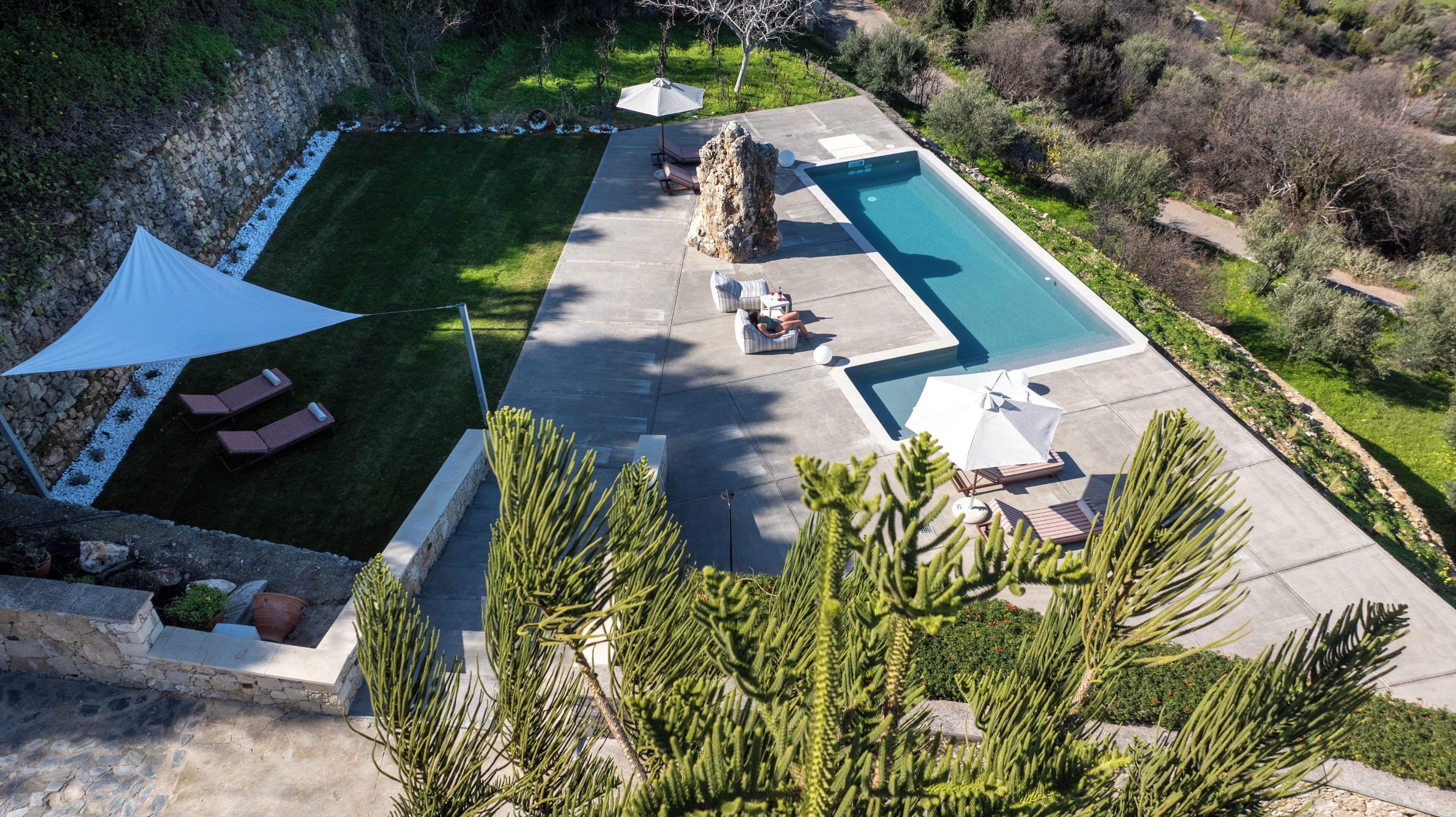 Vista aérea de un área de piscina privada con sillones de descanso, una gran formación rocosa y vegetación circundante.