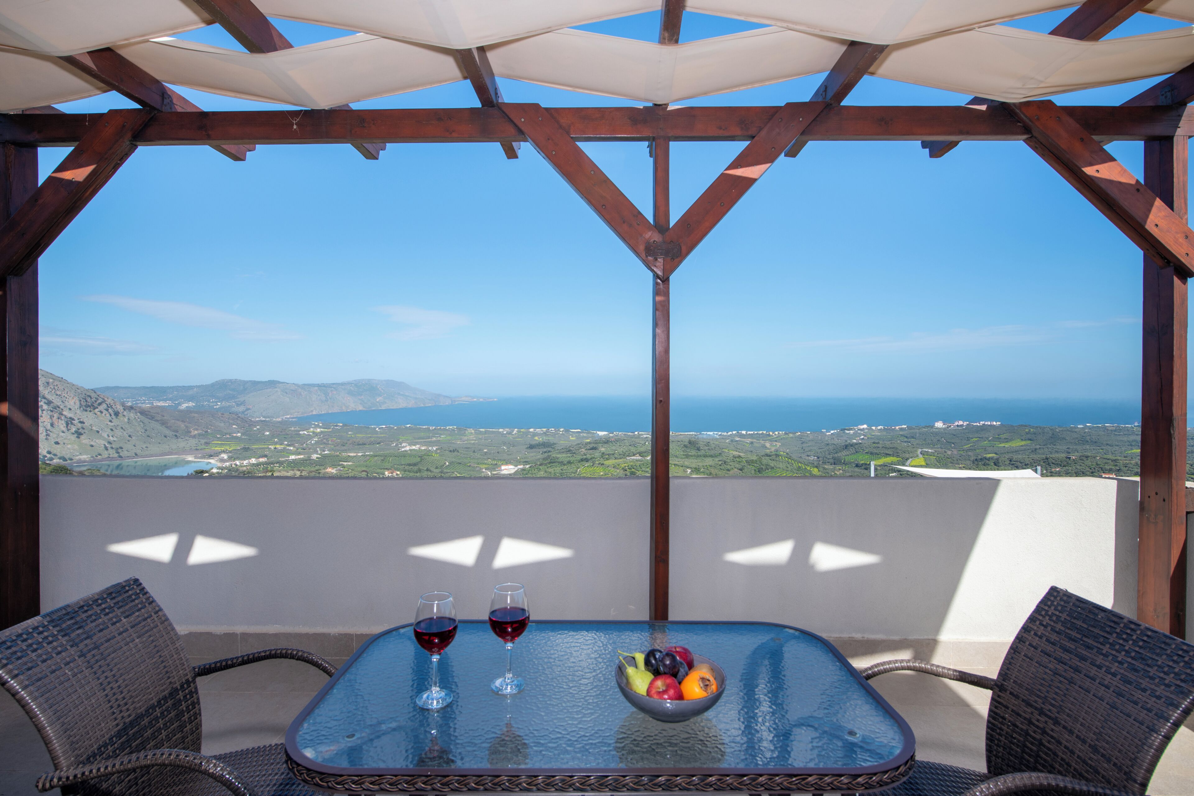 A balcony with a table, two glasses of red wine, and a bowl of fruit.