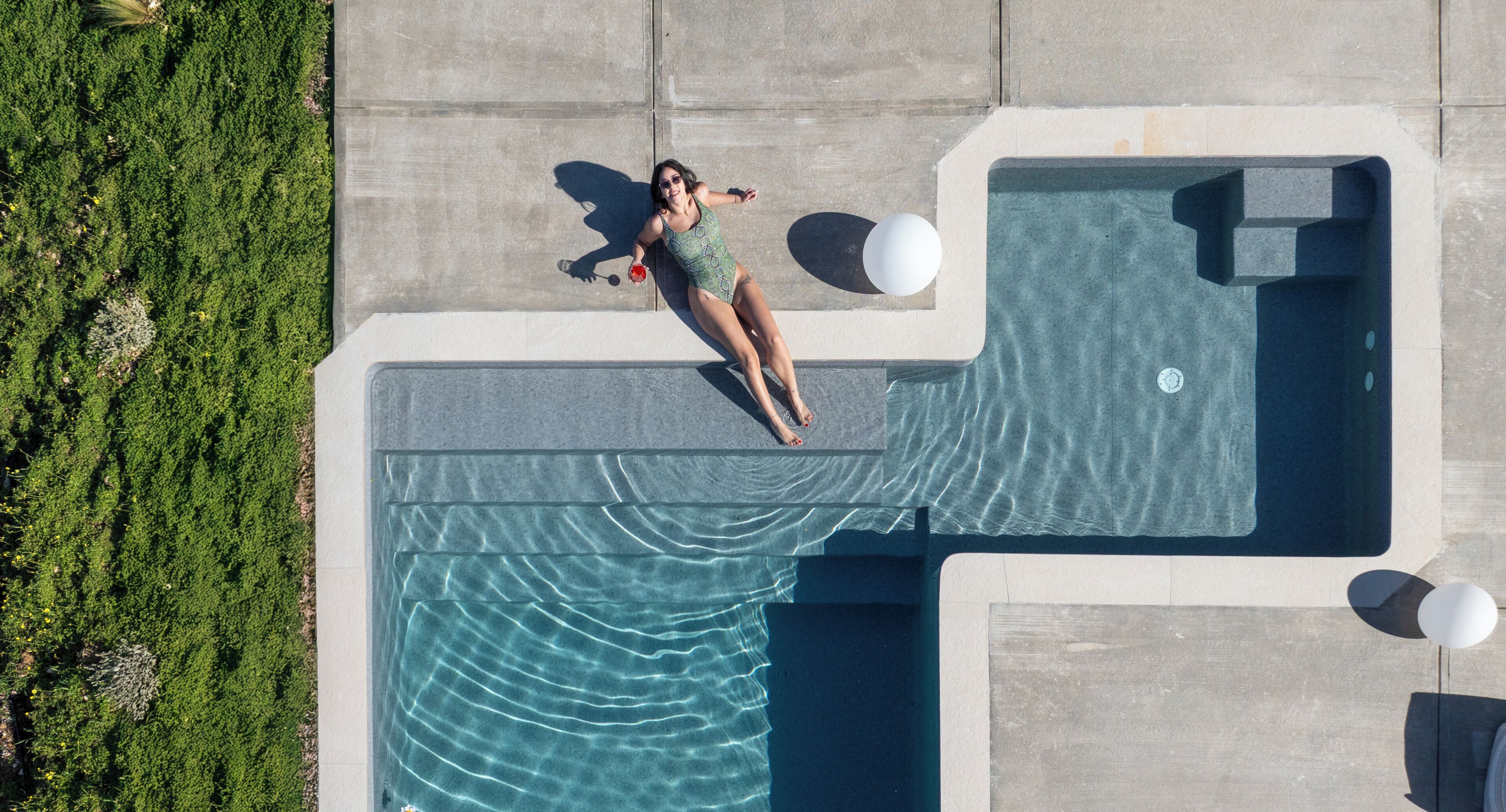 Vista aerea di una piscina con una persona sdraiata sul bordo, circondata da vegetazione e strutture in cemento.