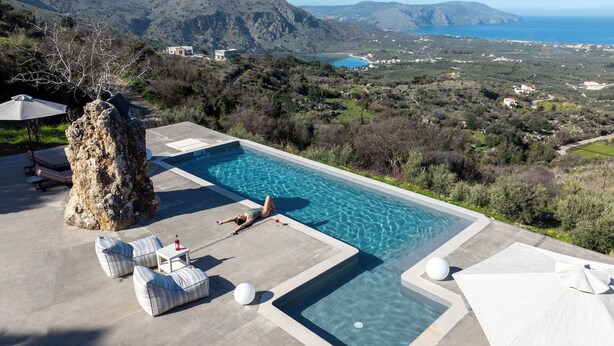 A pool with clear water, surrounded by lounge chairs and a stone pillar. - Thea Villas Complex (Apokoronas)