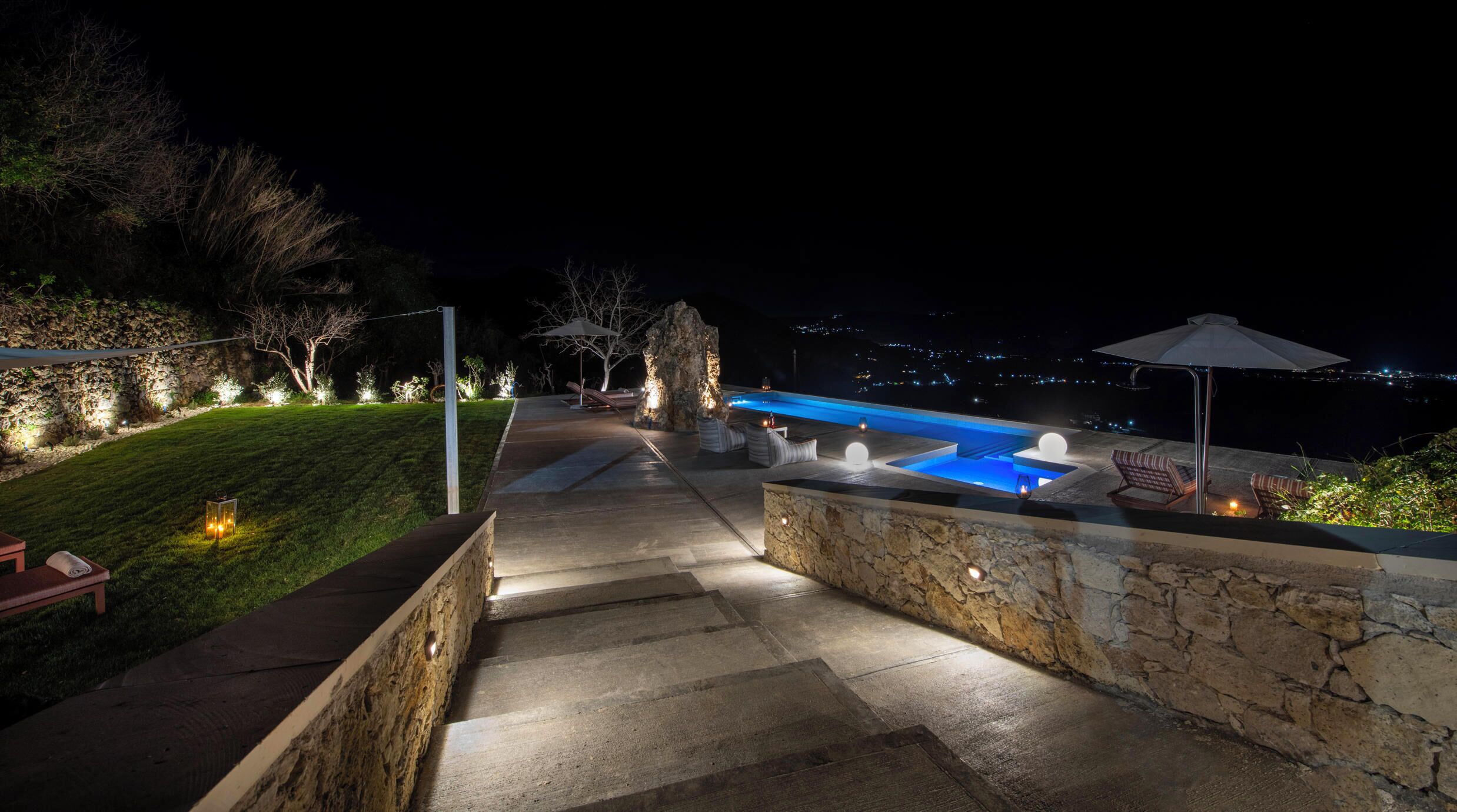 Ein Außenbereich bei Nacht mit Swimmingpool, Steinmauern und Blick auf die Stadt.