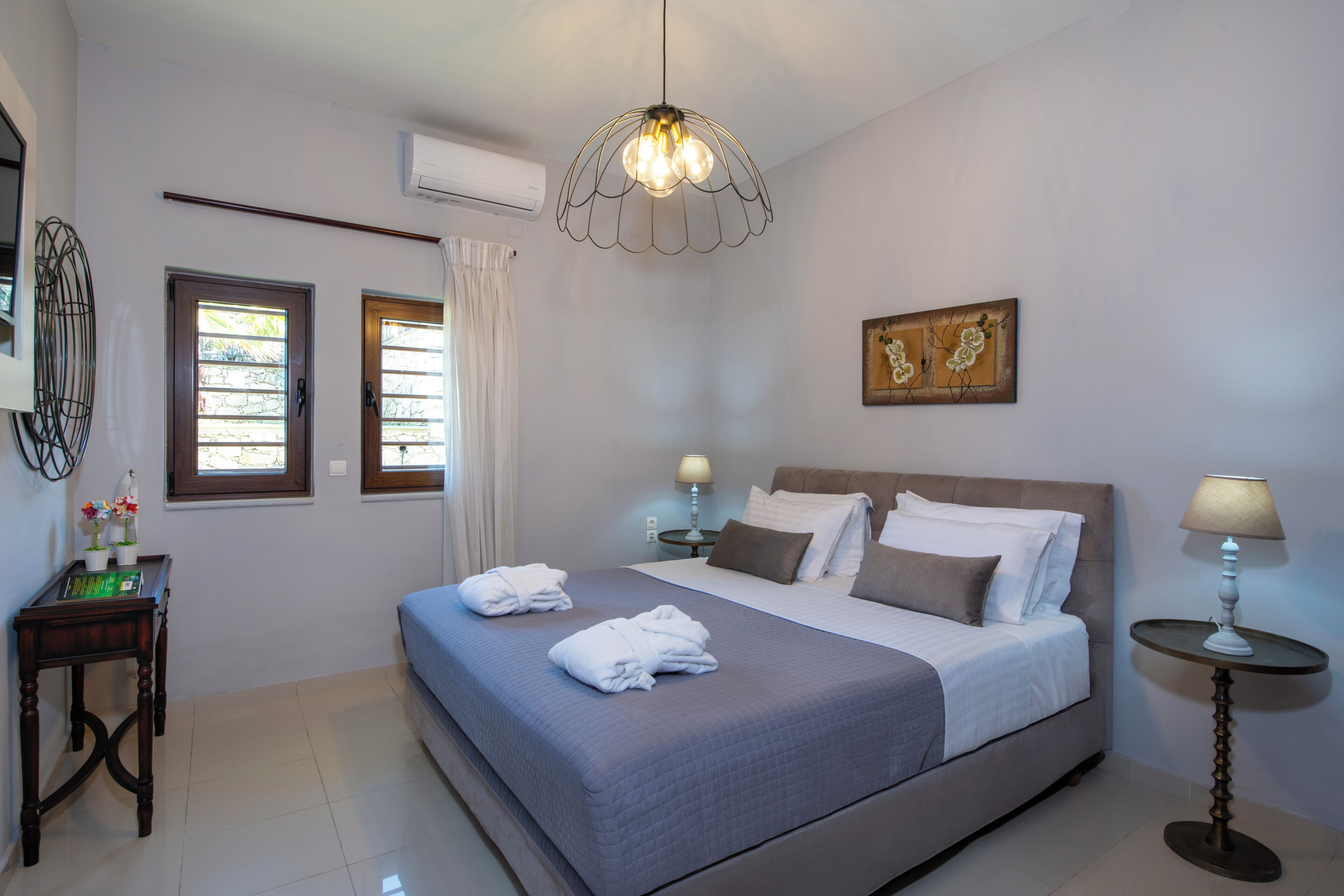 A bedroom with a bed, two bedside tables with lamps, a window with curtains, a wall-mounted air conditioner, and a decorative painting.