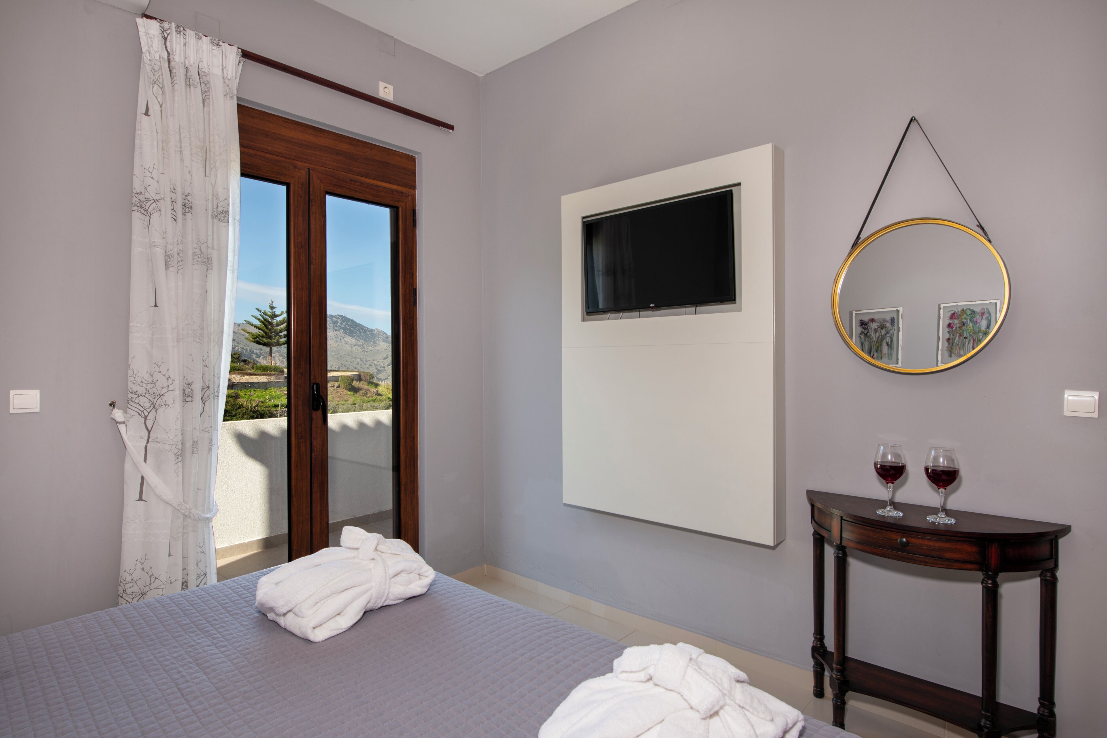 A bedroom with a bed, a bedside table with two glasses, a large window, and a wall-mounted TV.