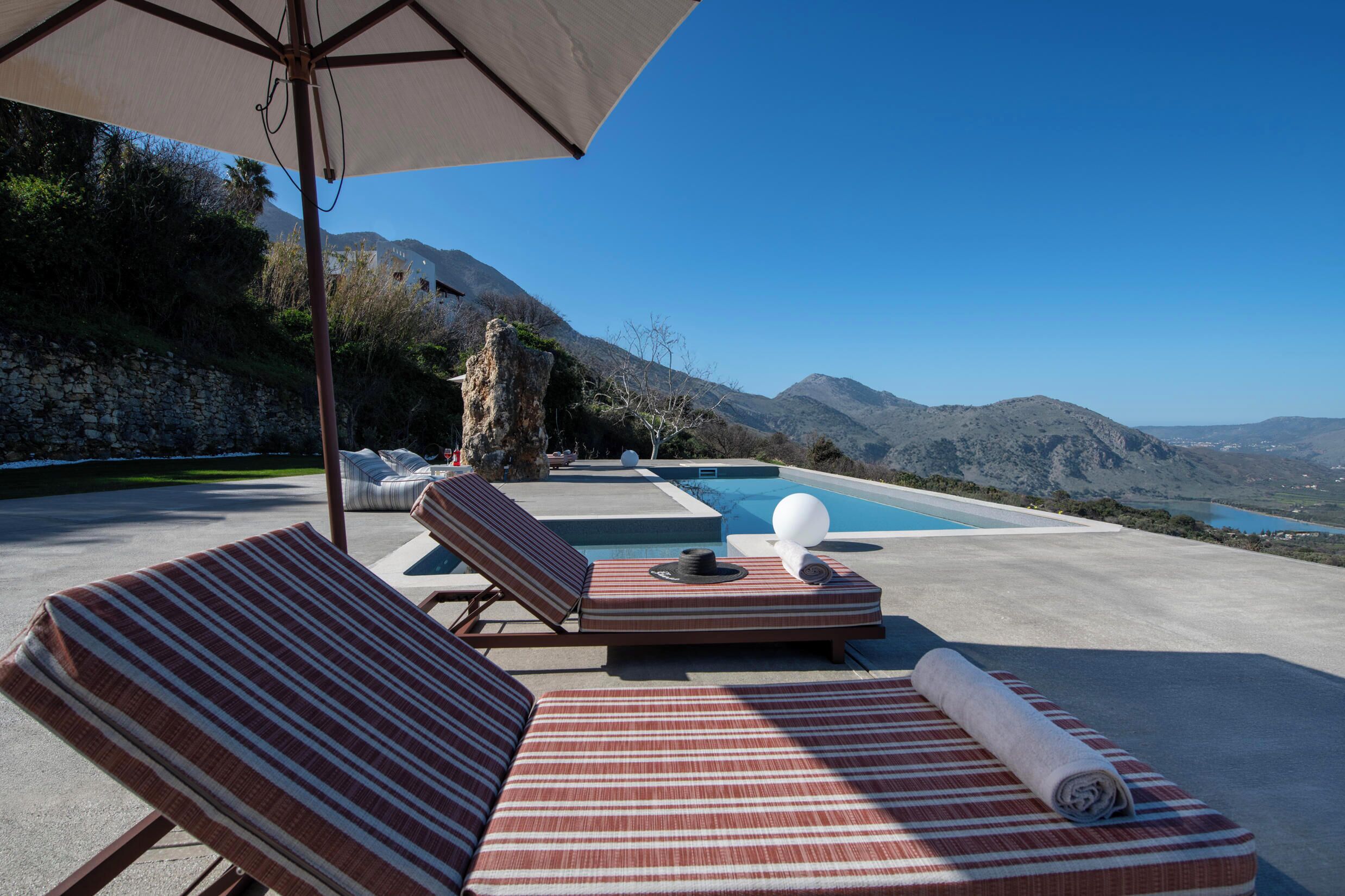 Zona de piscina con sillones de descanso y una sombrilla grande, con vista a un paisaje montañoso.