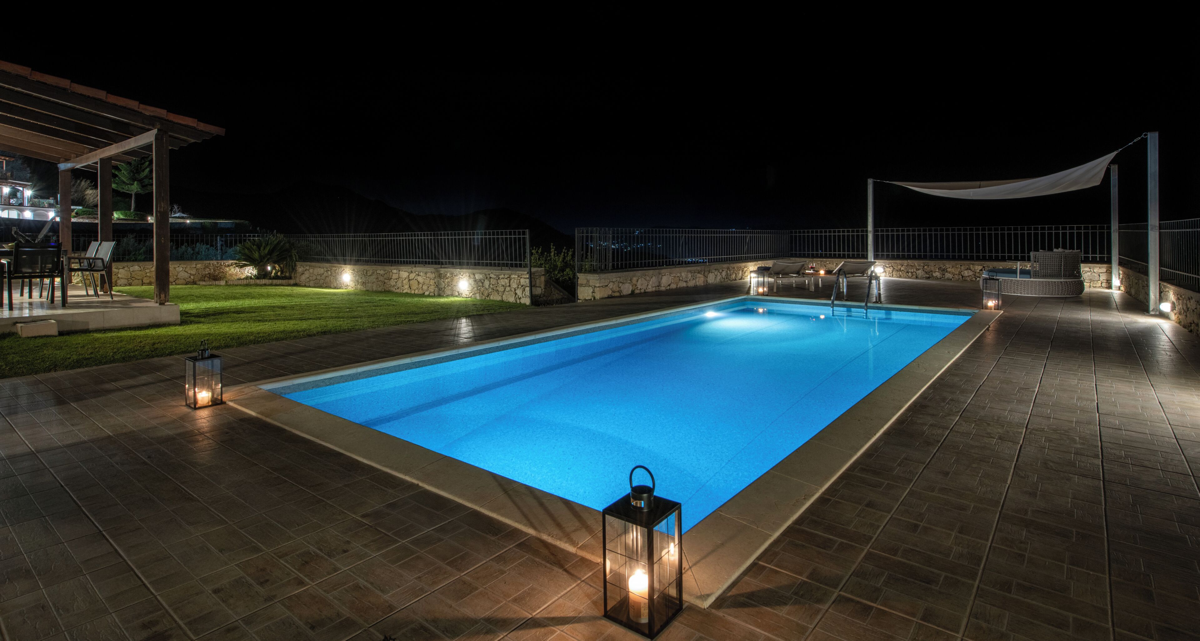 A rectangular swimming pool with a tiled deck, surrounded by stone walls and a white canopy.