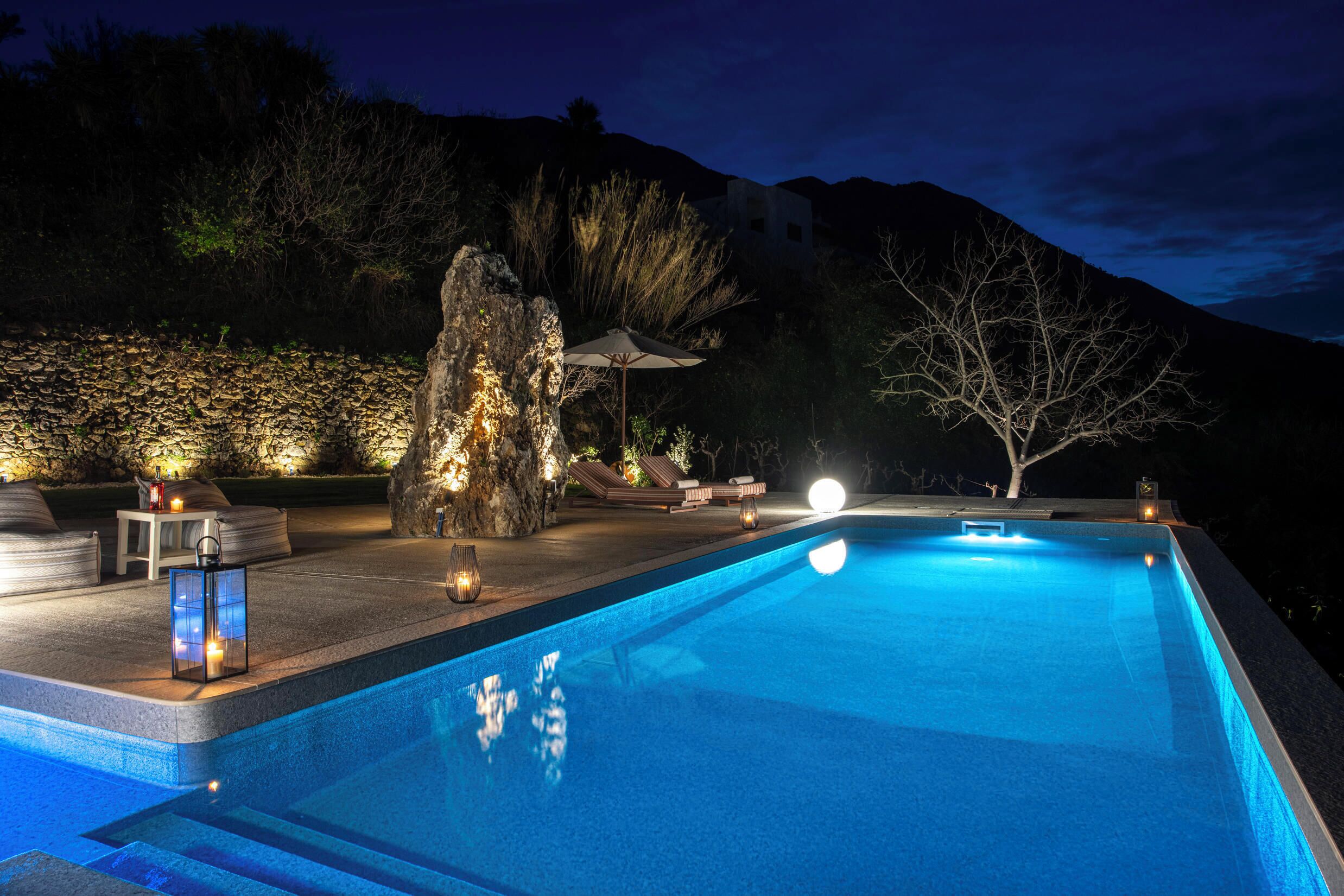 A swimming pool with a stone wall, lounge chairs, and a large rock centerpiece.