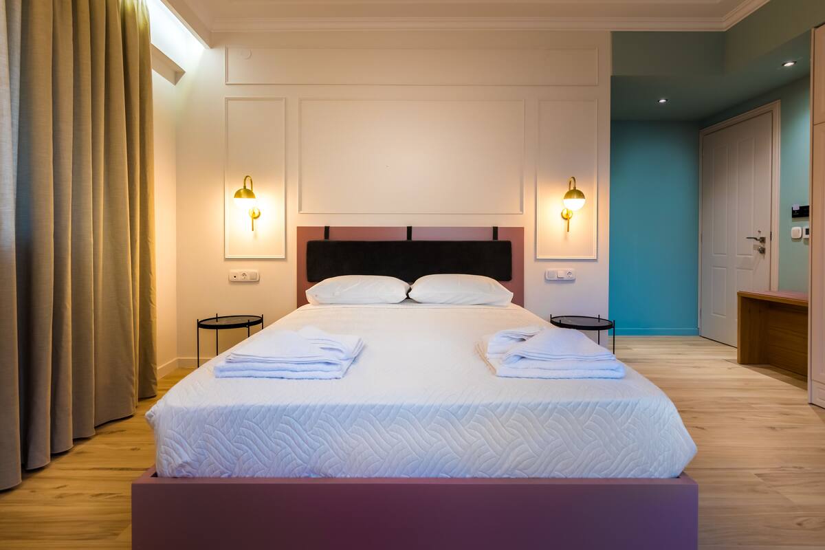 Una cama bien tendida con sábanas y almohadas blancas, dos mesitas de noche e iluminación instalada en la pared.