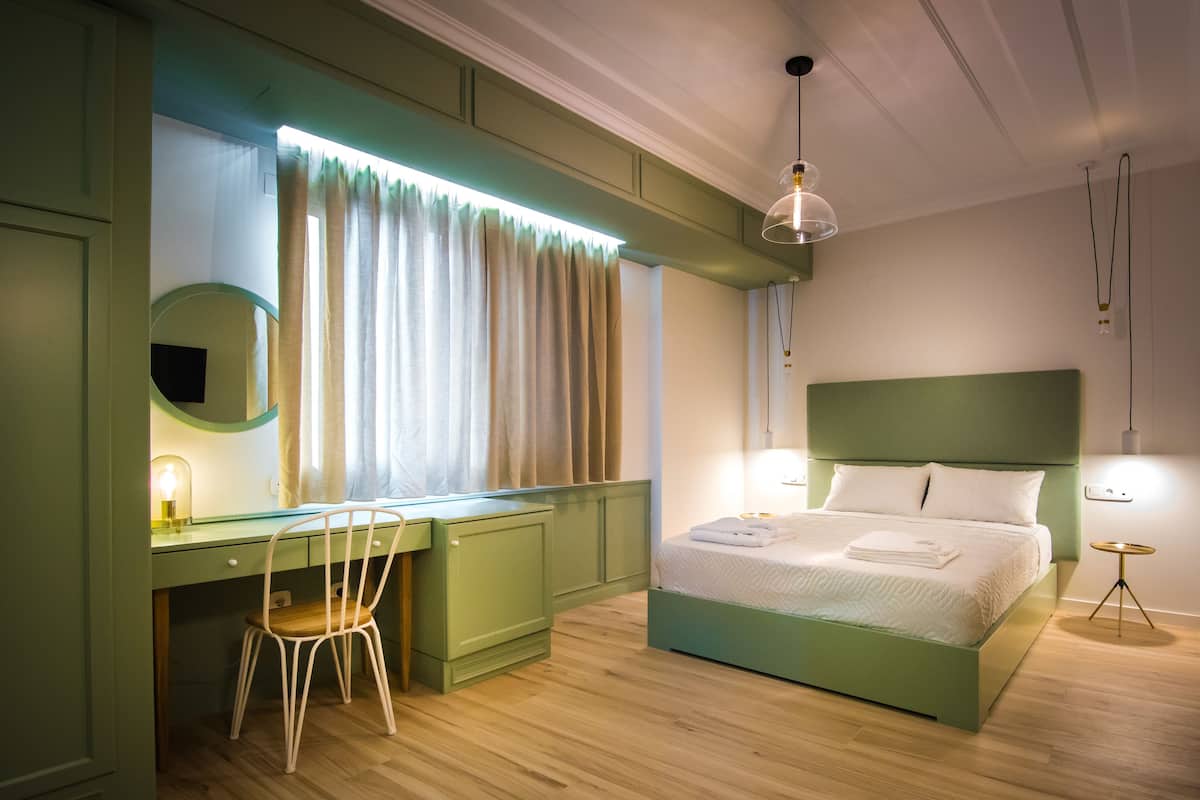 Un dormitorio con una cama verde, un escritorio con una silla, una ventana con cortinas y una lámpara colgante.