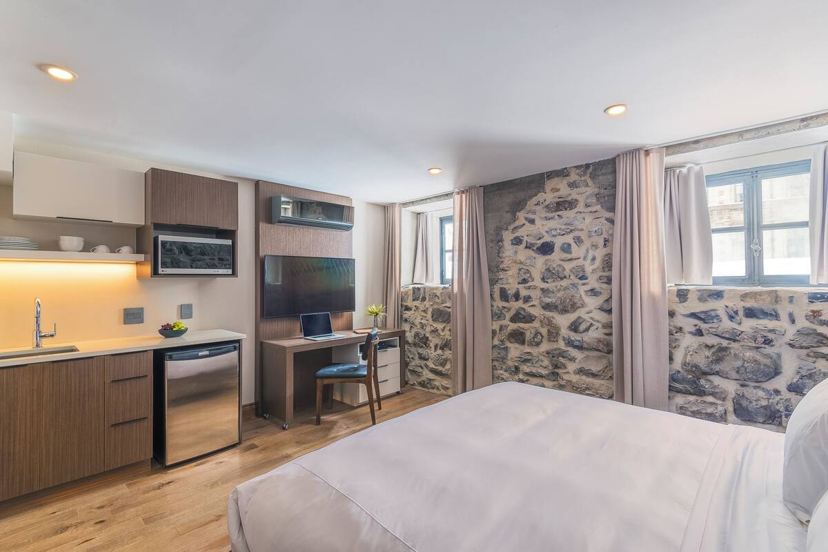 Habitación compacta con cama, escritorio con una computadora portátil, pequeña cocina y pared de piedra.