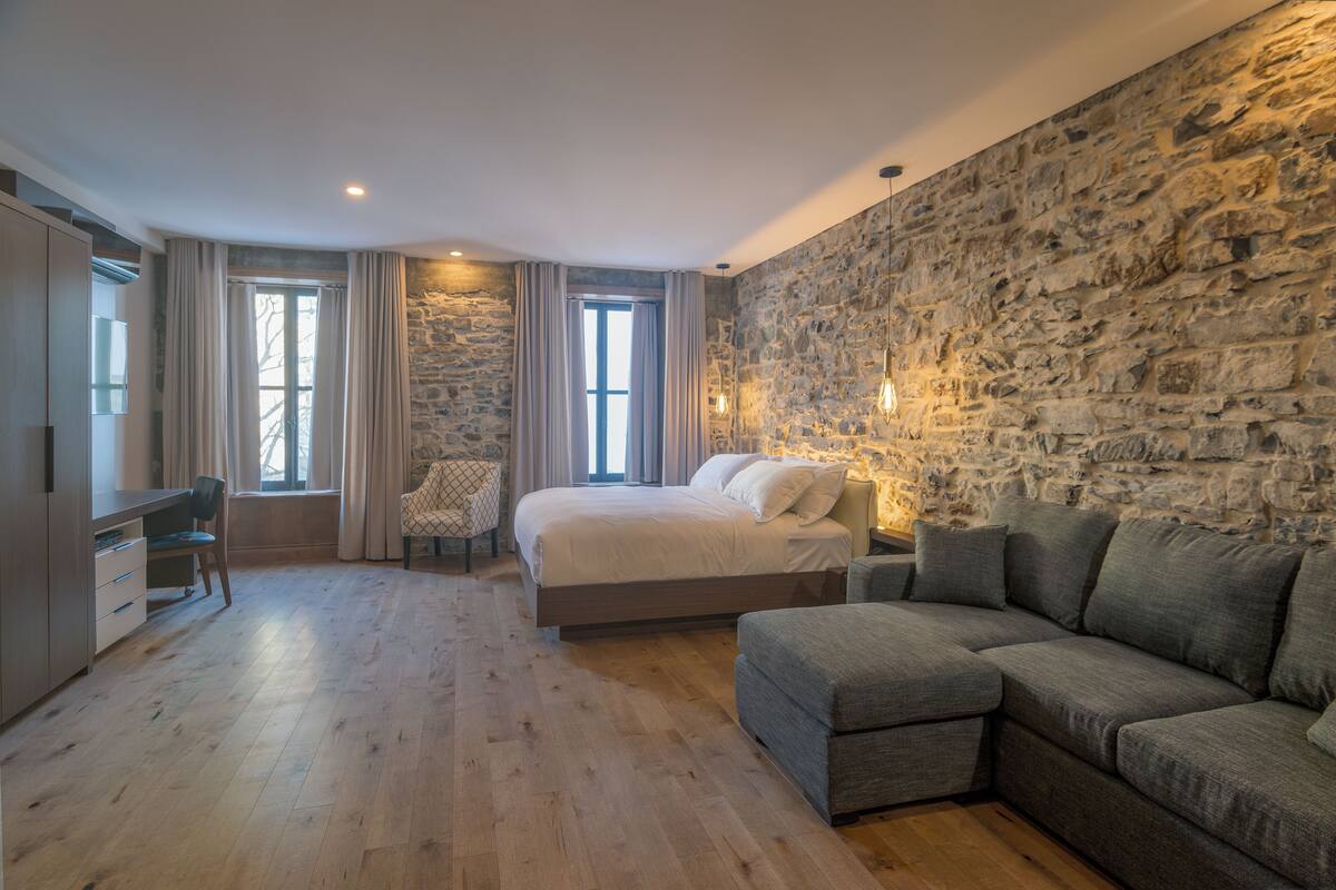 Un dormitorio moderno con pared de piedra, una cama grande, un sofá gris y un escritorio con una silla.