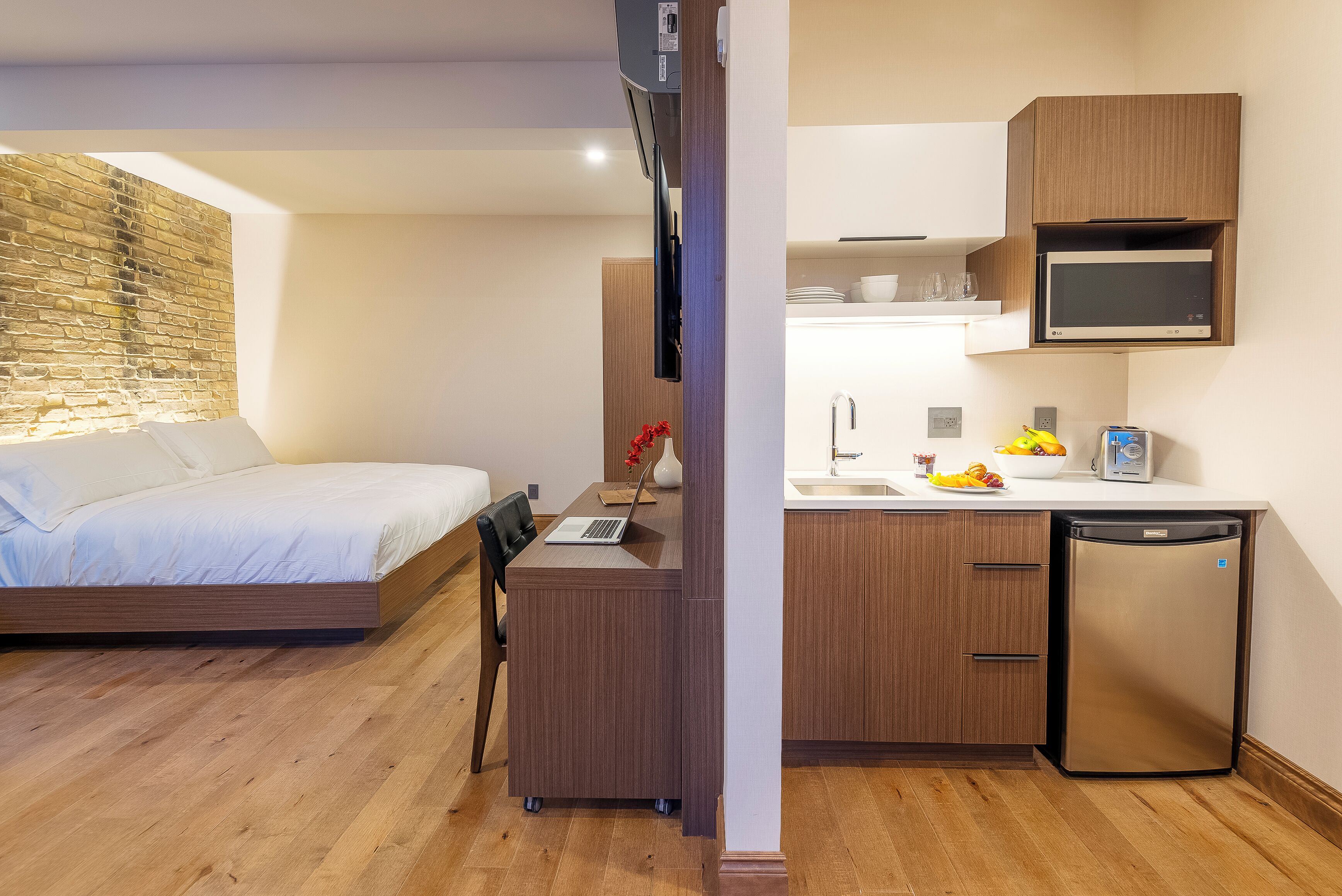 A compact hotel room with a bed, a desk with a laptop, a small kitchen area with a microwave, and a dishwasher.