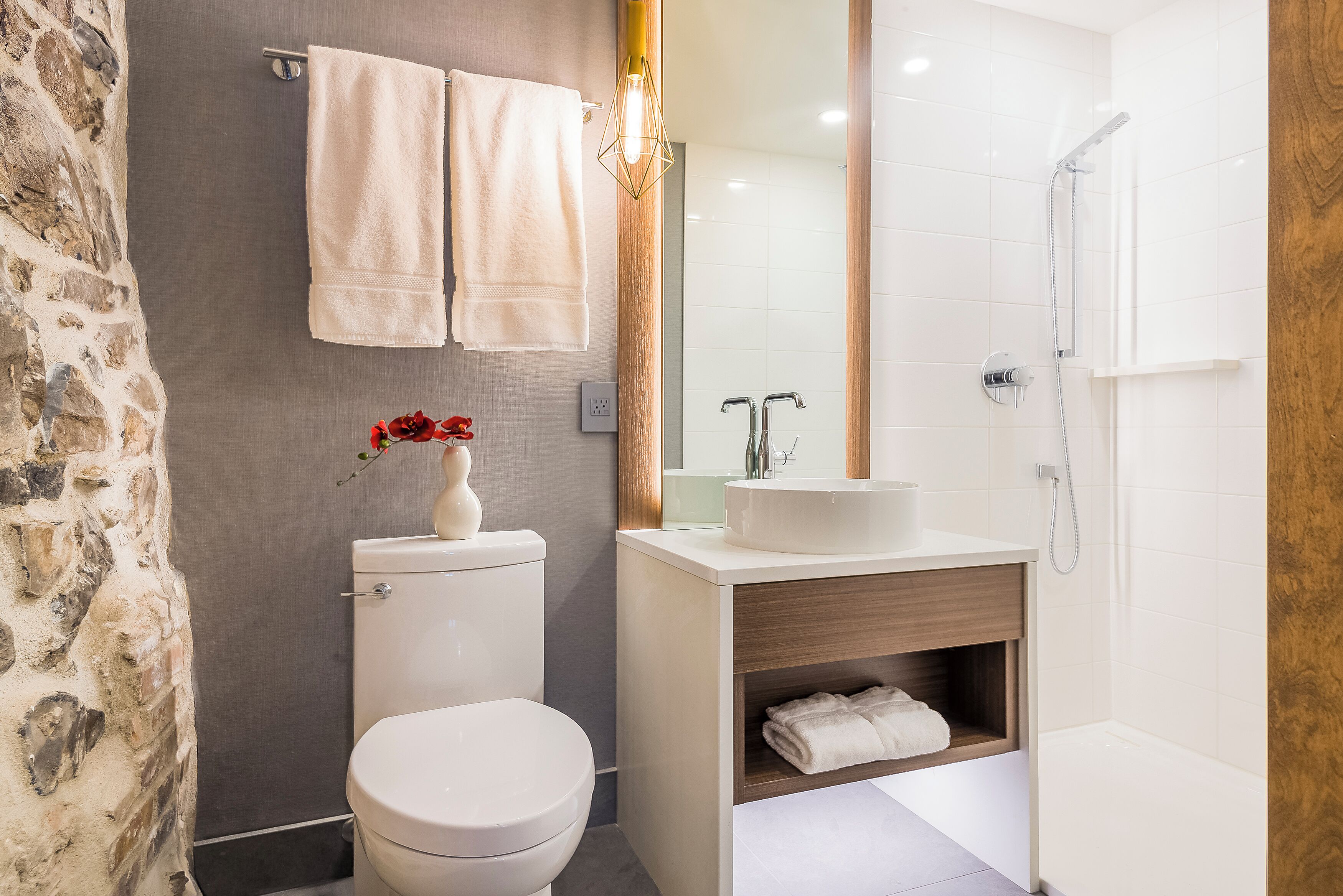 Un baño con inodoro, lavamanos y ducha. Hay toallas colgadas en la pared y un florero con flores sobre el lavamanos.