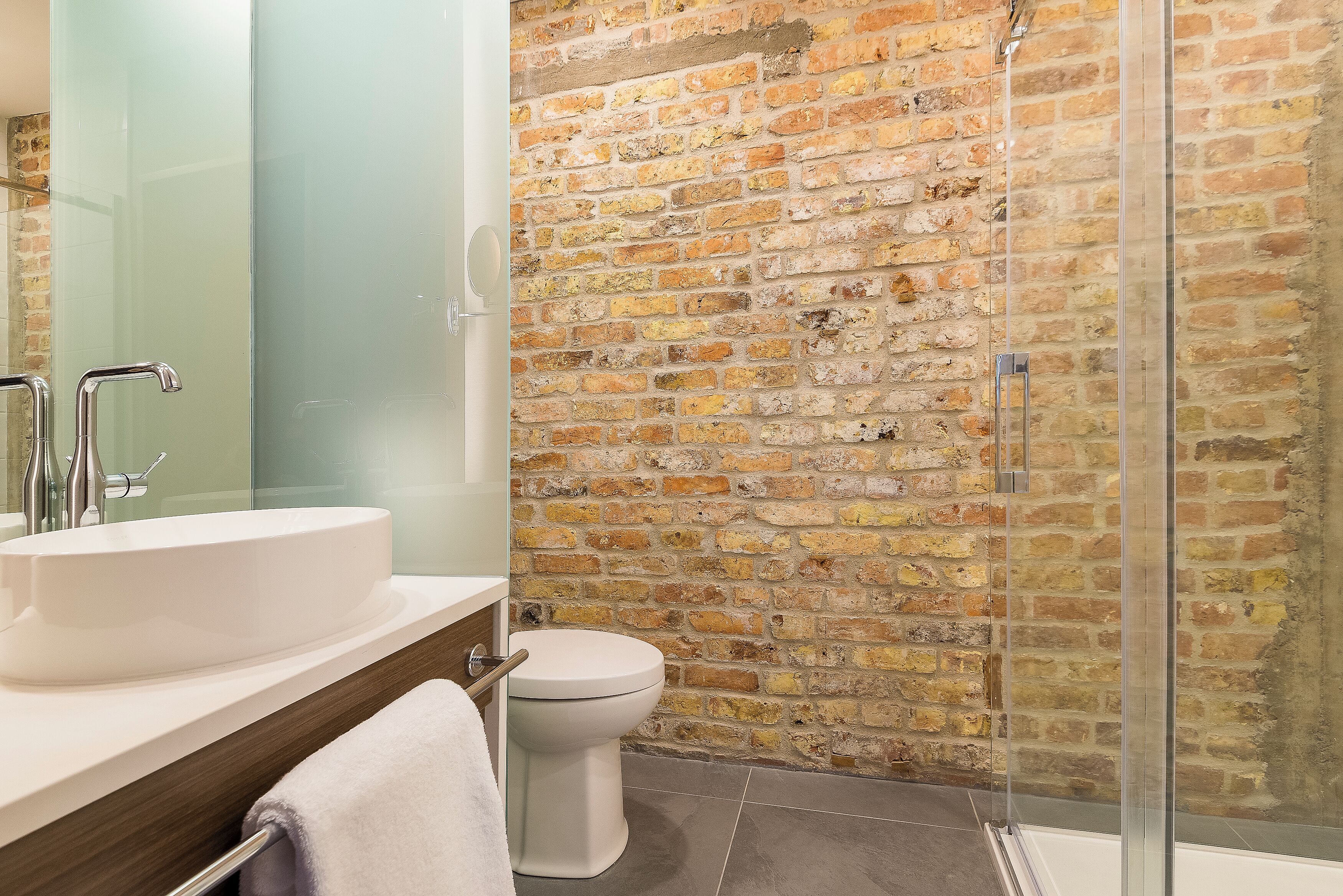A bathroom with a white sink, a toilet, and a glass shower enclosure with a brick wall.