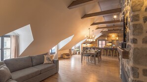 A loft-style living area with a high ceiling, exposed beams, a stone wall, a dining area with stools, and a kitchen in the background. - La Maison Kent - Par Aneyro (Québec City)