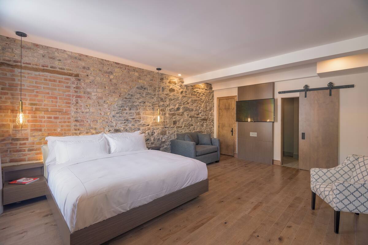 Un dormitorio con pared de ladrillo, una cama, un sillón gris, un armario de madera y un televisor fijado a la pared.